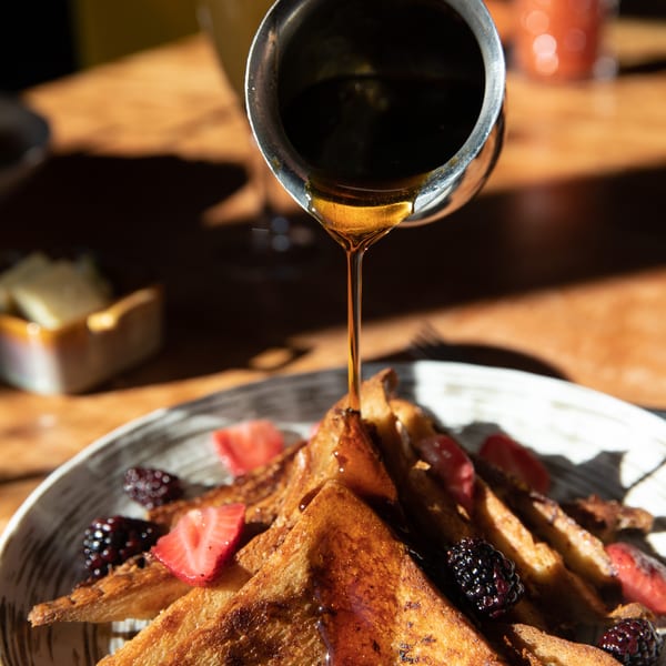 Maple syrup pouring over golden French toast with fresh strawberries, blueberries, and blackberries.