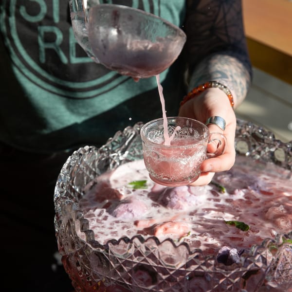 A person with tattooed arms ladles pink punch from a large crystal bowl into a small glass.