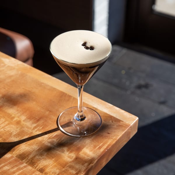 Espresso martini with foam and three coffee beans on a wooden table in sunlight.