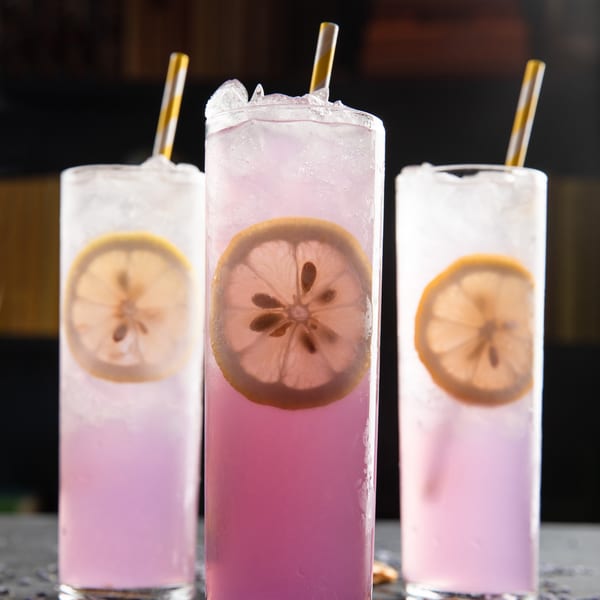 Three tall glasses of purple lemonade with lemon slices, ice, and striped straws.