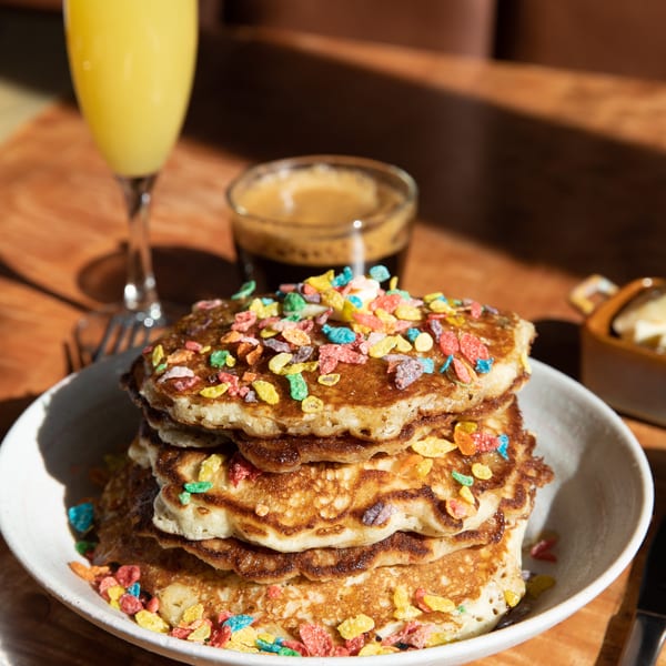 Stack of pancakes topped with colorful cereal, alongside a mimosa and espresso on a wooden table.
